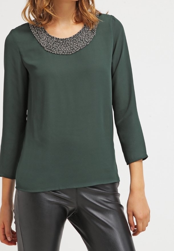 Blouse verte à manches longues avec un col rond orné de perles noires et argentées, associée à un pantalon en cuir noir. Texture du tissu lisse.