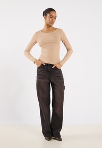 Langermet, ribbet, lysebrun topp med rund hals, kombinert med løse, mørkeblå jeans. Modellen står med hendene i lommene.