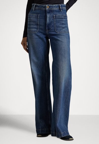 Forfra af mørkeblå vidde jeans med store forlommer båret af en person iført sort top og sorte spidse sko.