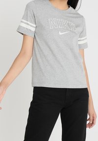 Camiseta gris de manga corta con el logo "NIKE" en blanco en el pecho y dos rayas blancas en cada manga. Fabricada con una suave mezcla de algodón.