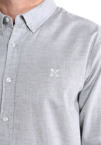 Chemise boutonnière gris clair en tissu avec une finition légèrement texturée. Dotée d'un col et d'un petit motif brodé blanc sur la poitrine.