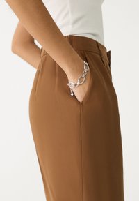 Brown tailored trousers with a smooth texture, featuring a side pocket. A silver chain bracelet is visible on the wrist.