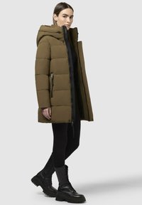 Oliven grøn pufferjakke med hætte, med lynlås lukning og sidelommer. Tekstureret materiale med quiltet design, parret med sorte støvler.