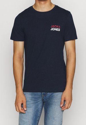 Mężczyzna ubrany w czarny t-shirt z krótkim rękawem z logo "JACK & JONES" w kolorze czerwonym i białym na piersi, w połączeniu z niebieskimi dżinsami.