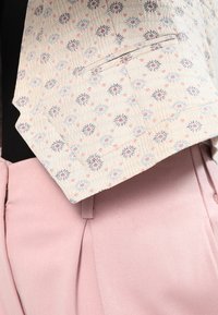 Béžová bunda s malým květinovým potiskem, texturovaná tkanina a jedna kapsa, spárovaná se světle růžovými kalhotami s ostrými záhyby.