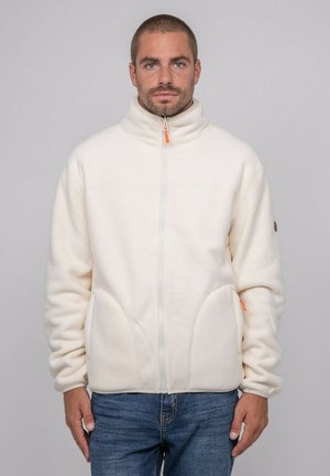 Homme portant une veste polaire crème zippée à col montant et un jean bleu, debout devant un fond uni gris clair.