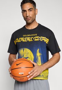 Μαύρο μπλουζάκι με το κείμενο «Golden State Warriors» σε κίτρινο, με γραφικό ενός μπάσκετ και μπλε λεπτομέρειες. Παίκτης που κρατά μια πορτοκαλί μπάλα του μπάσκετ.