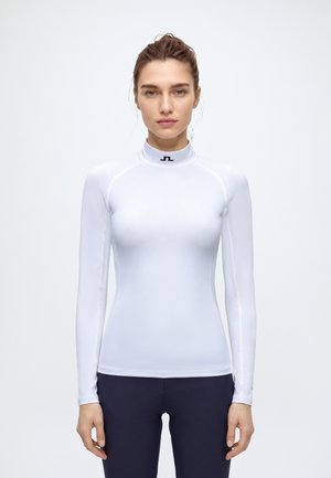 Une femme porte un haut blanc ajusté à manches longues et à col montant, orné d’un petit logo noir, ainsi qu’un pantalon foncé, sur un fond uni.