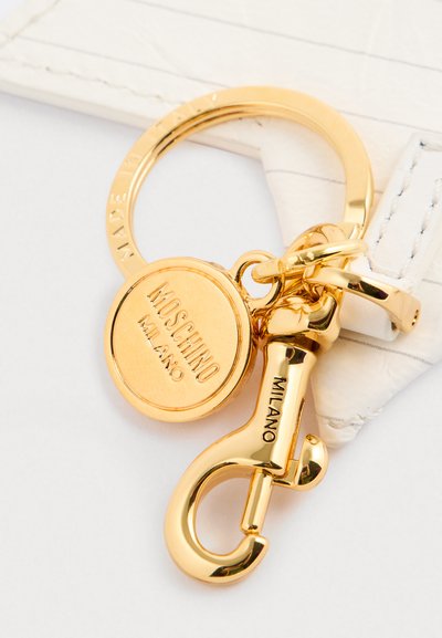 Złoty breloczek z okrągłym zawieszką z wytłoczonym napisem "Moschino Milano" oraz dekoracyjnym zapięciem w kształcie haka. Gładkie wykończenie metalowe, wypolerowany tekstura.