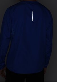 Chemise de sport à manches longues en tissu bleu avec une bande verticale réfléchissante dans le dos. Texture lisse et coupe décontractée.