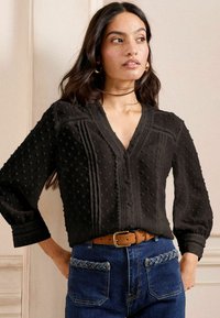 Blusa nera con pois testurizzati, scollatura a V e chiusura con bottoni. Presenta cuciture dettagliate e maniche lunghe rilassate. Indossata con jeans blu.