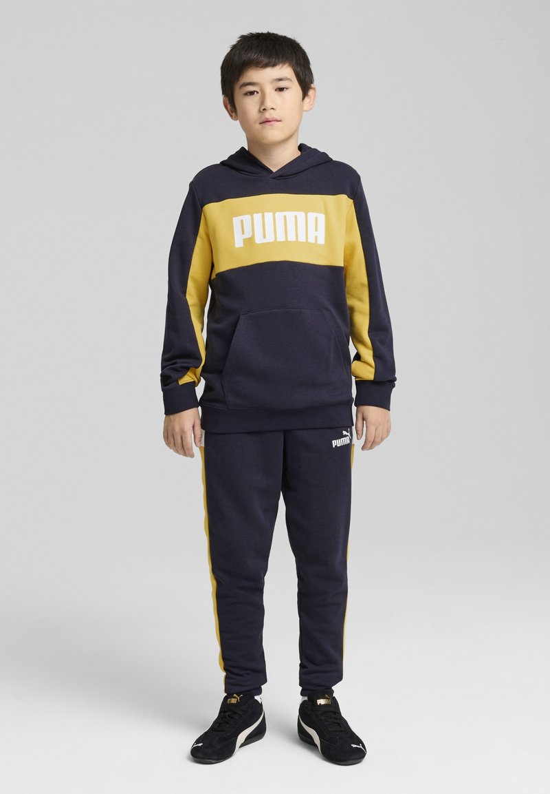 Granatowa bluza z kapturem z żółtą linią i białym logo "PUMA"; w komplecie z dopasowanymi granatowymi spodniami z żółtymi paskami po bokach i logo.