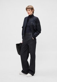 Melna bomber jaka, tumšs augsts apkakle džemperis, plašas tumšas džinsa bikses, baltas sporta apavi un melna tote soma. Teksturēti materiāli; brīvs piegriezums.