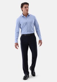 Camicia azzurra chiara con bottoni e maniche lunghe, abbinata a pantaloni scuri e scarpe nere. Tessuto liscio con vestibilità classica e leggero lucido.