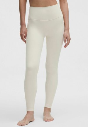 Kobieta w beżowych, wysokich, długich legginsach stojąca boso na jednolitym tle.