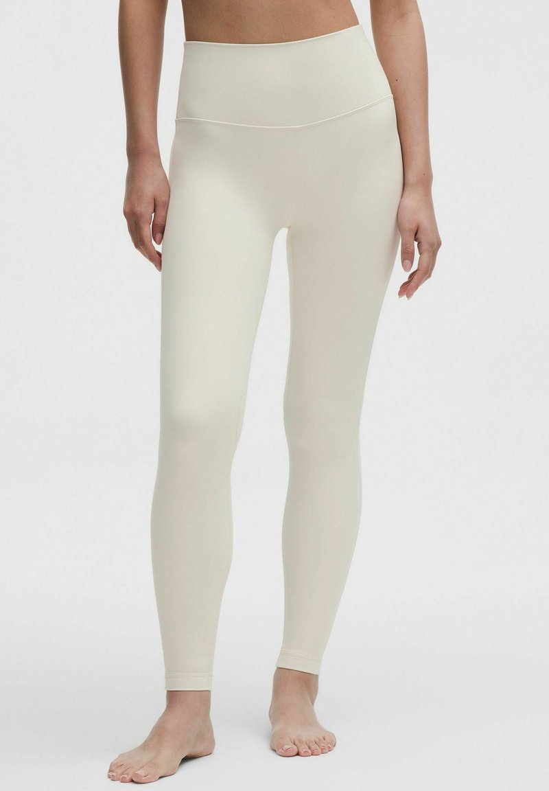 Kobieta w beżowych, wysokich, długich legginsach stojąca boso na jednolitym tle.