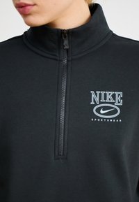 Melnā Nike sporta ceturtdaļas apģērba vējdrošā augstajā apkaklē, ar baltu Nike logotipu un "SPORTA APĢĒRBS" tekstu. Gluda auduma tekstūra.
