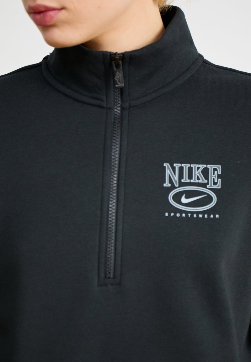 Camiseta deportiva negra de Nike de media cremallera con un alto collar, que presenta un logotipo blanco de Nike y el texto "SPORTSWEAR". Textura de tejido suave.