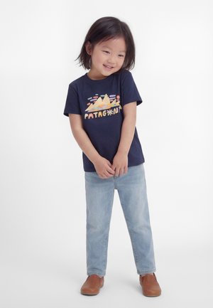 Jong kind met kort donker haar dat lacht, draagt een marineblauw T-shirt met een afbeelding, lichtblauwe spijkerbroek en bruine schoenen, staat in een ontspannen houding.