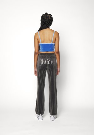 Frau mit geflochtenem Haar steht mit dem Rücken zur Kamera, trägt ein blaues Crop-Top, schwarze Velourhosen mit dem Text "Juicy" und weiße Turnschuhe.