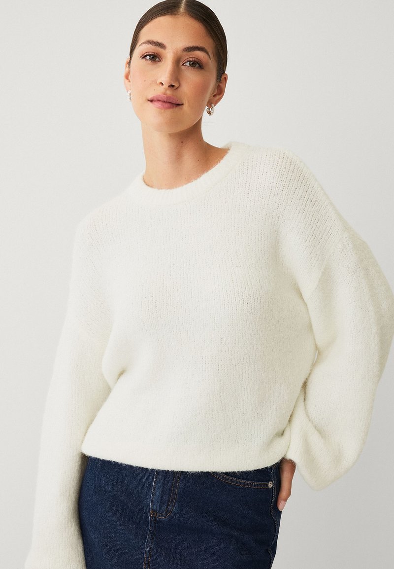 Pull en tricot blanc et moelleux avec un col rond et des épaules tombantes ; associé à un denim foncé, mettant en valeur une coupe décontractée et une texture douce.