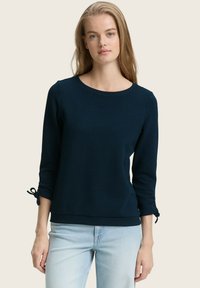 Donna con lunghi capelli biondi che indossa un top a maniche lunghe blu scuro con polsini annodati e jeans blu chiaro, in piedi su uno sfondo neutro.