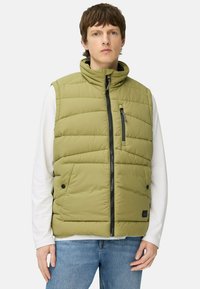 Padded olivgrüwest mit Stehkragen, vorne mit Reißverschluss, zwei seitlichen Taschen und einer strukturierten Oberfläche. Getragen über einem weißen Langarmshirt.