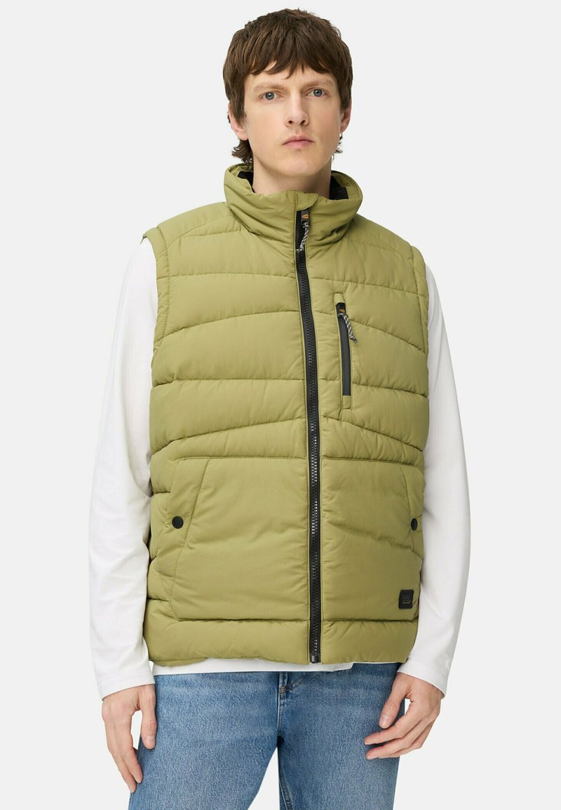 Padded olivgrüwest mit Stehkragen, vorne mit Reißverschluss, zwei seitlichen Taschen und einer strukturierten Oberfläche. Getragen über einem weißen Langarmshirt.