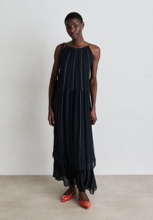 Dark pleated maxi dress with a high neckline, thin straps, layered design, and contrasting light stripes; paired with bright red flats.