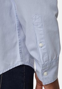 Camicia azzurro chiaro a maniche lunghe con polsini abbottonati, design delle maniche a punta e dettagli di cucitura puliti. Il tessuto appare liscio e simile al cotone.