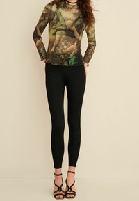 Gennemsigtig langærmet top med et farverigt blomstermotiv, parret med sorte leggings og højhælede sandaler med stropper.
