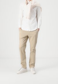 Beżowe chinosy, biała, długosleeve'owa koszula z guzikami i kremowy sweter w warkocze zarzucony na ramiona. Białe sneakersy dopełniają stylizację.