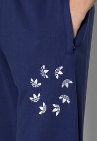 En persons hand i fickan på marinblå byxor med vita Adidas trefoil-logotyper arrangerade i ett cirkulärt mönster på låret.