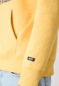 Sweatshirt jaune en tissu doux, avec une poche kangourou et des poignets côtelés. Petite étiquette logo GANT sur la manche.