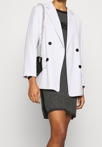 Blazer gris clair croisé à boutons noirs, porté sur une robe noire et argentée à texture, assorti d'un petit sac à main noir.