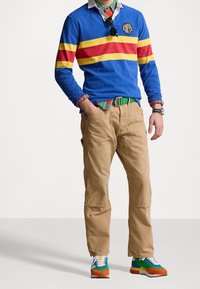 Blå långärmad polo med röda och gula ränder, khakifärgade byxor, grön bälte och färgglada sneakers med orange, blå och gröna accenter.