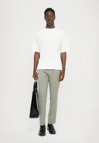 White short-sleeve t-shirt paired with light grey trousers. Holding a black textured tote bag. Black loafers complete the outfit.