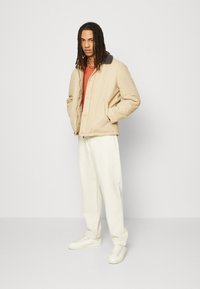 Beige jacket with a dark collar, featuring a front closure and side pockets, paired with white pants and white sneakers.