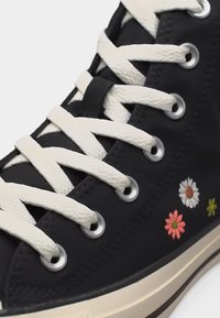 Μαύρο αθλητικό παπούτσι υψηλού πάνω μέρους με λευκά κορδόνια και κεντητά ροζ, λευκά και κίτρινα λουλούδια κοντά στη σόλα.