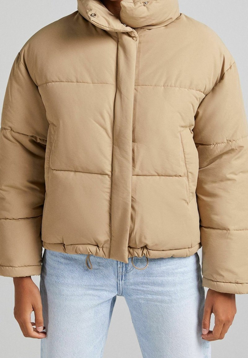 Giacca puffer beige con colletto alto, chiusura a snap e coulisse elastica all'orlo. Tessuto testurizzato e vestibilità ampia e oversize.