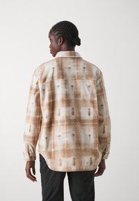 Fleece skjorte i brune og kremfargede tie-dye mønster, med klassisk krage, knappebeslåtte ermer, og forlenget bakre søm. Myk tekstur.