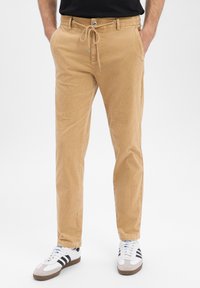 Beżowe sztruksowe spodnie ze ściągaczem w pasie i dwoma bocznymi kieszeniami, o gładkiej fakturze i zwężanym kroju. Połączone z białymi i czarnymi sneakersami.