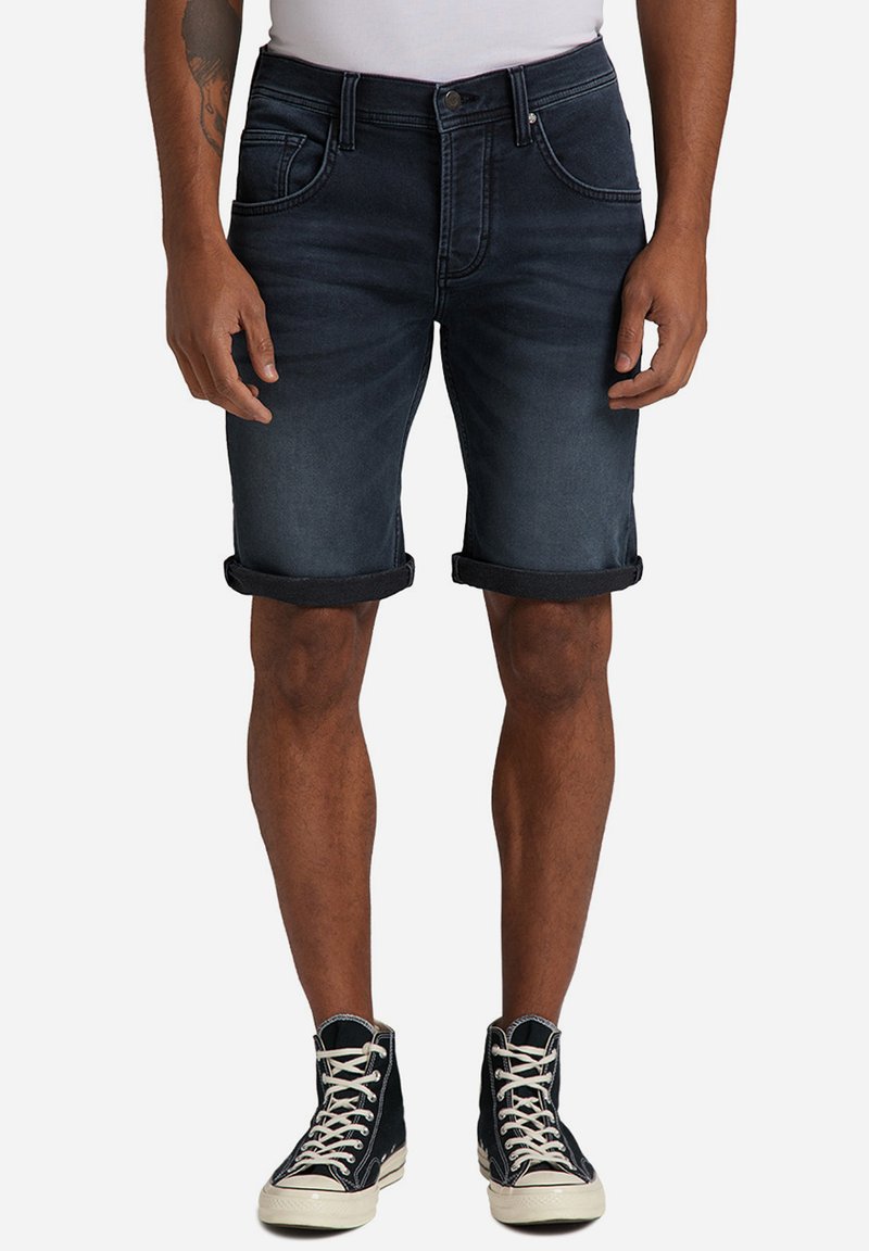 Pantalones cortos de mezclilla en azul oscuro con dobladillos enrollados, con bolsillos frontales y un cierre de botón simple. Combinados con zapatillas negras de caña alta.