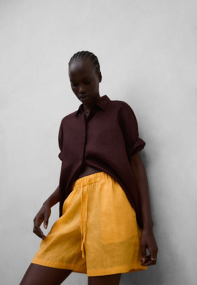 Femme aux cheveux tressés portant une chemise marron foncé fluide et un short à cordon de serrage jaune vif, debout devant un fond gris uni.