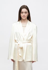 Kremo spalvos satino švarkas su surišama juosmens detale, ilgomis rankovėmis ir dviem priekinėmis kišenėmis. Lygios tekstūros, su struktūruotais pečiais ir nertos apykaklės dizainu.