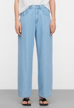 Persoon draagt lichtblauwe jeans met wijde pijpen, witte ingestopte top en zwarte open-toe sandalen, staand op een grijze vloer met een effen witte achtergrond.