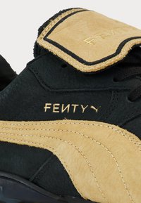 Black suede sneaker featuring a tan leather accent, textured material, prominent FENTY branding, and a distinctive silhouette with a padded tongue.