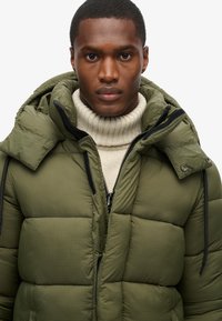 Oliven grøn pufferjakke med hætte, der har et rutenetmønster, lynlås og sorte detaljer. Båret over en cremefarvet turtleneck.