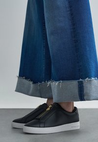 Ténis de couro preto com fecho de correr e detalhes dourados, combinados com culotes de ganga azul com bainhas desfiadas e punhos cinza claro.