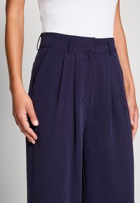 Marineblauwe geplooide broek met een gladde textuur, voorzien van een rechte pasvorm, steekzakken aan de zijkant en een op maat gemaakte tailleband.